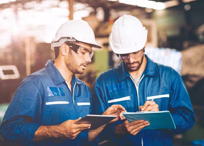 Deux ingénieurs en casque et lunettes analysant des données sur tablette en atelier industriel.