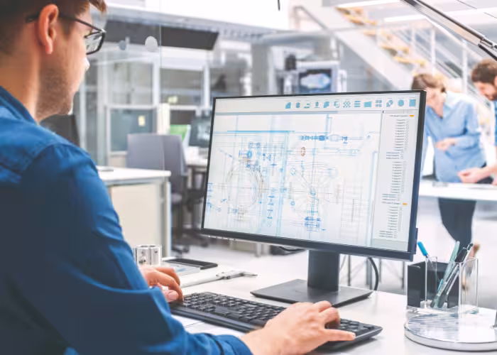 Ingénieur devant écran affichant plan technique détaillé, collègues discutant en arrière-plan.