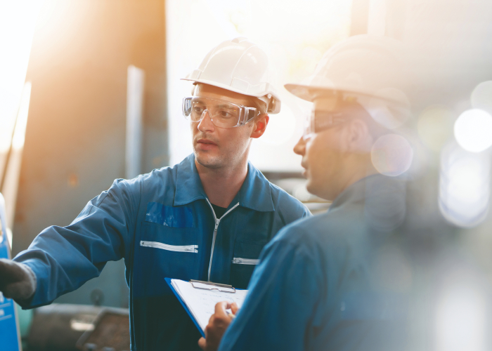 Deux techniciens en casque et lunettes inspectant un équipement industriel avec tablette et dossier.