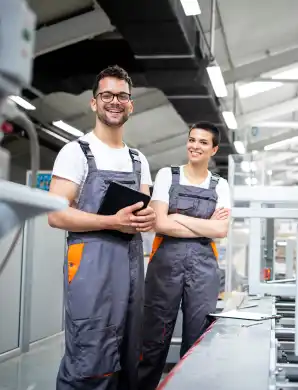 Deux opérateurs souriants en salopette tenant une tablette dans un atelier industriel moderne.